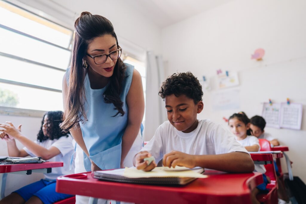 Public school teacher helping a young student at a classroom desk.