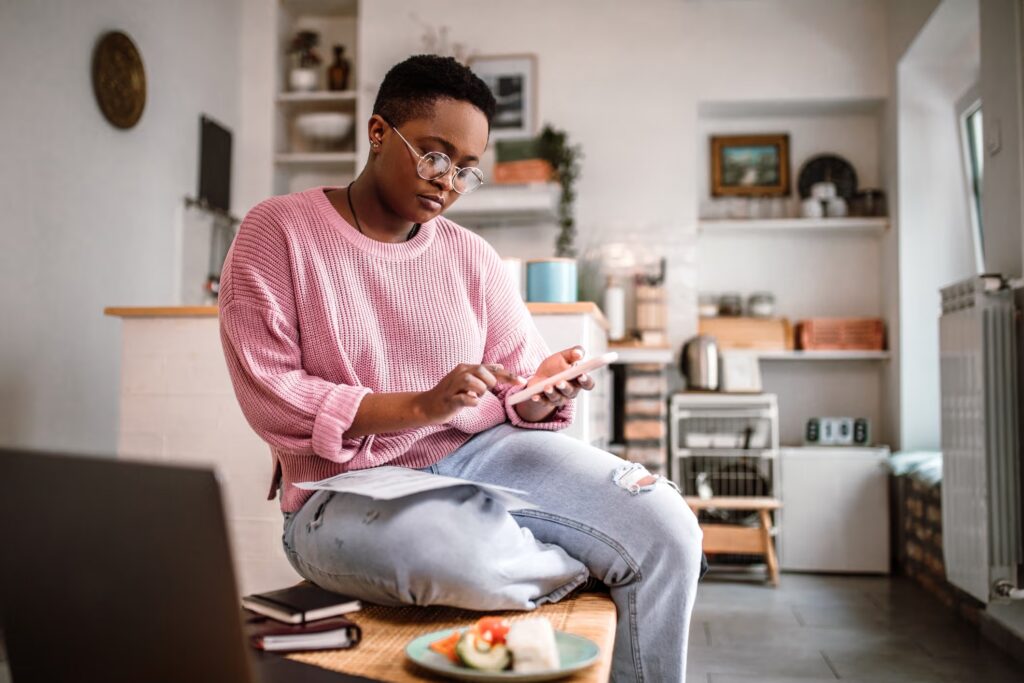 A woman reviewing information on her phone at home, with a laptop open in front of her.