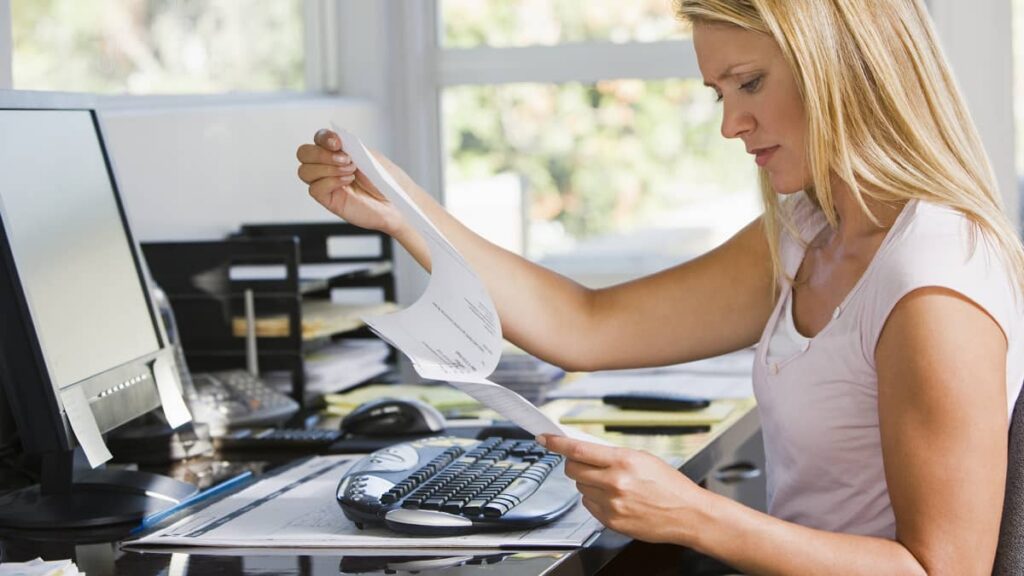 A borrower at a home desk reading a printed student loan statement beside a computer.
